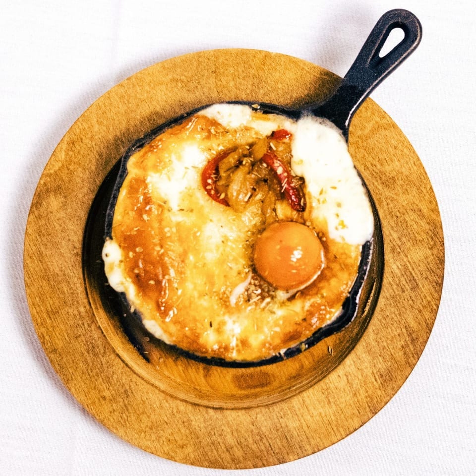 A sizzling skillet of baked cheese with a sunny-side-up egg and vegetables on a wooden plate.