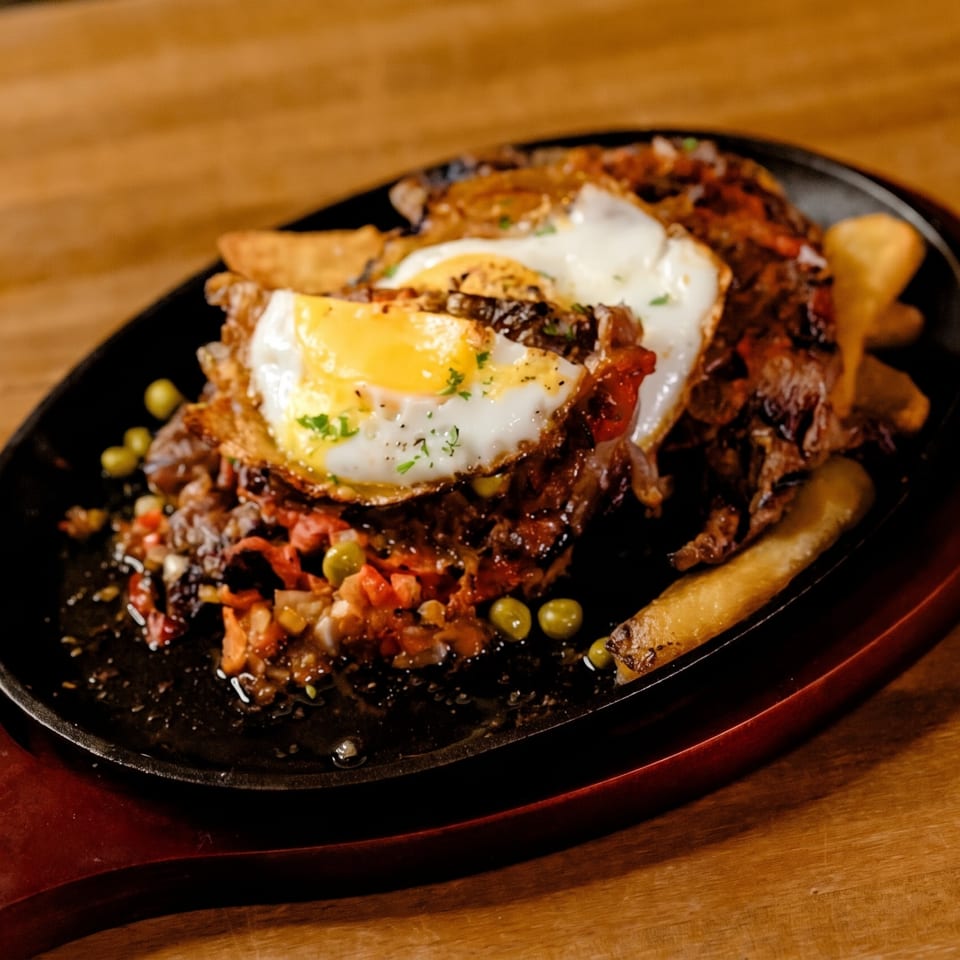 Sizzling meat dish with two fried eggs, vegetables, and fries on a black skillet.