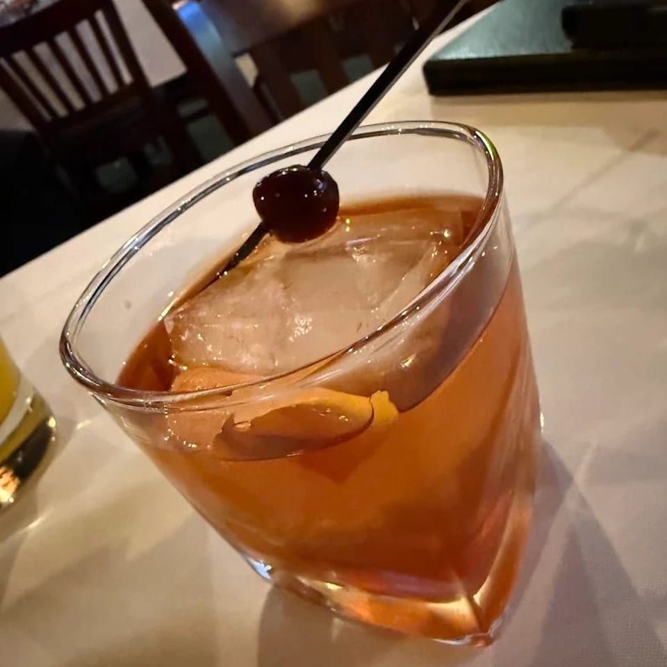 An Old Fashioned cocktail with a cherry and orange peel on a white tablecloth in a restaurant.
